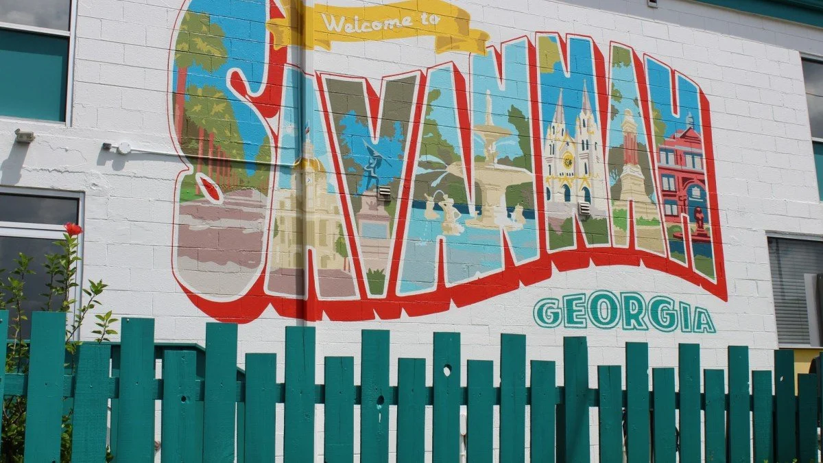 Colorful mural on a white brick wall welcoming visitors to Savannah, Georgia, with illustrations of landmarks and a teal fence in the foreground.