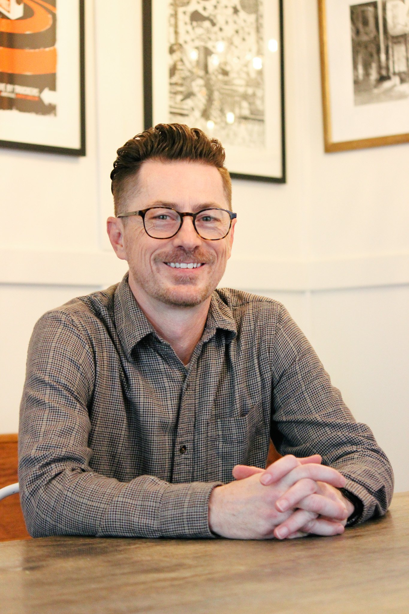 A man with glasses and short brown hair, smiling, sitting at a table with his hands clasped in front of him, in a room decorated with black and white framed photographs on the wall behind him.