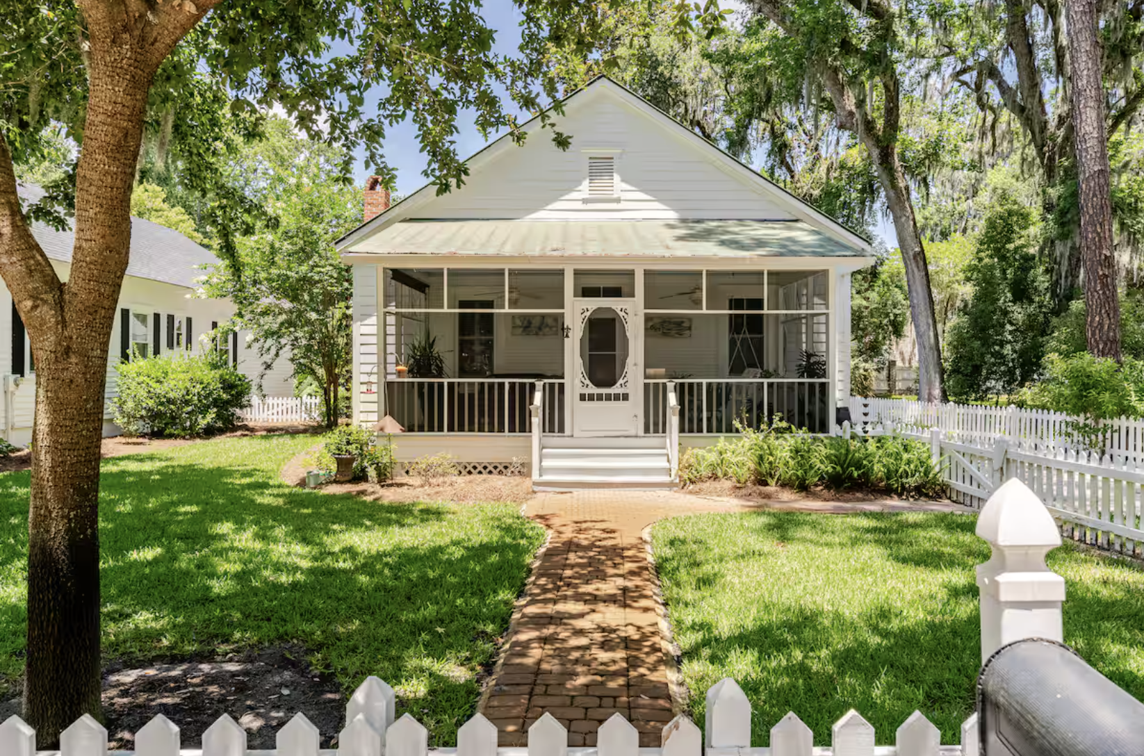 White house with a front porch, surrounded by a green lawn and tall trees, fenced with a white picket fence and a brick pathway leading to the front steps.