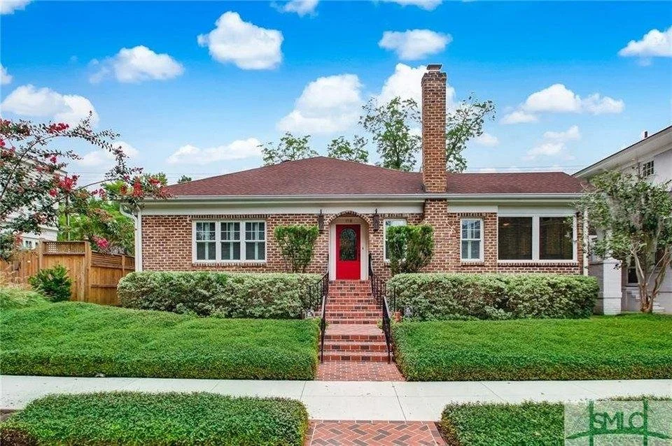 Brick house with a red door, central staircase, and prominent chimney surrounded by a well-manicured lawn and bushes, under a partly cloudy sky.
