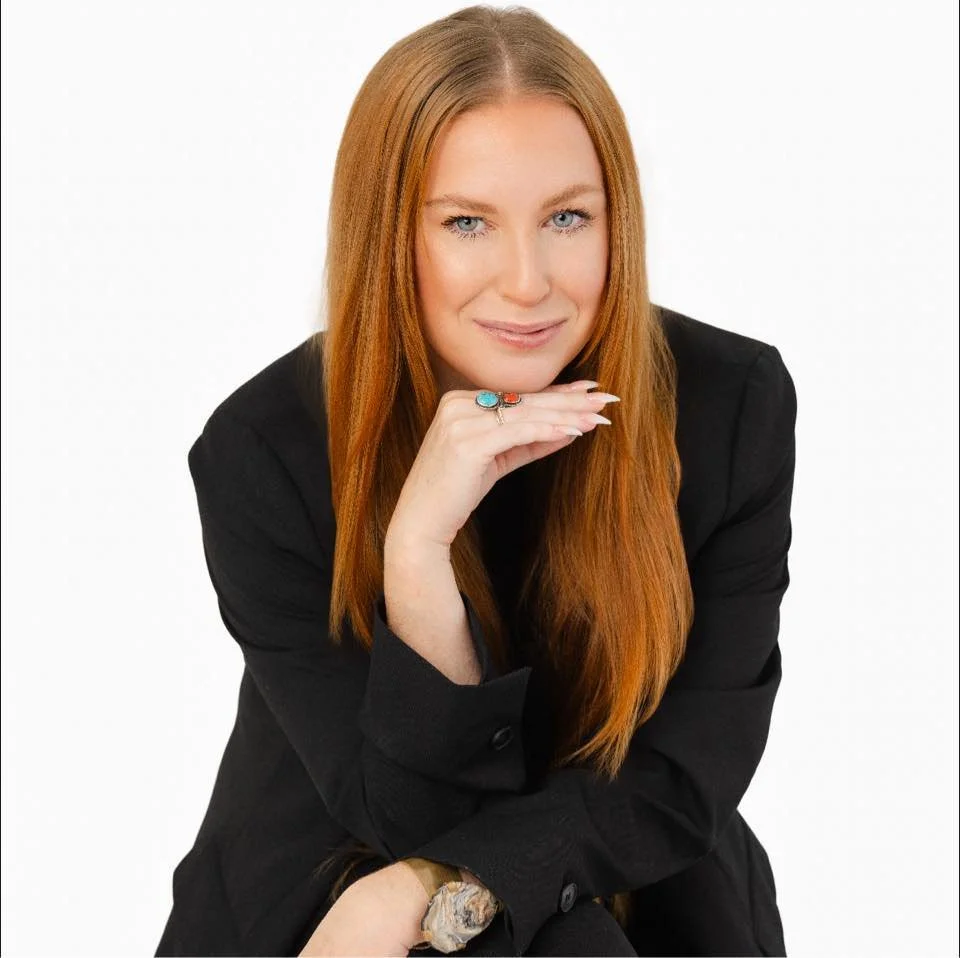 A woman with long red hair wearing a black blazer, sitting with her chin resting on her hand, and smiling at the camera against a white background.