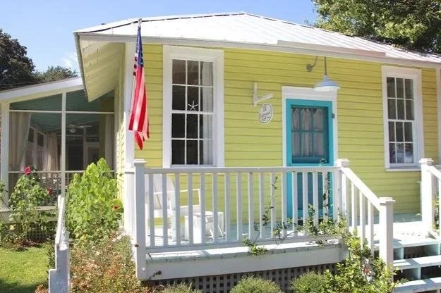 A small yellow house with white trim, a blue door, and a porch with white railings. An American flag hangs from the porch, and there's a large window on either side of the door. A clock is mounted on the wall near the door, and greenery surrounds the house.