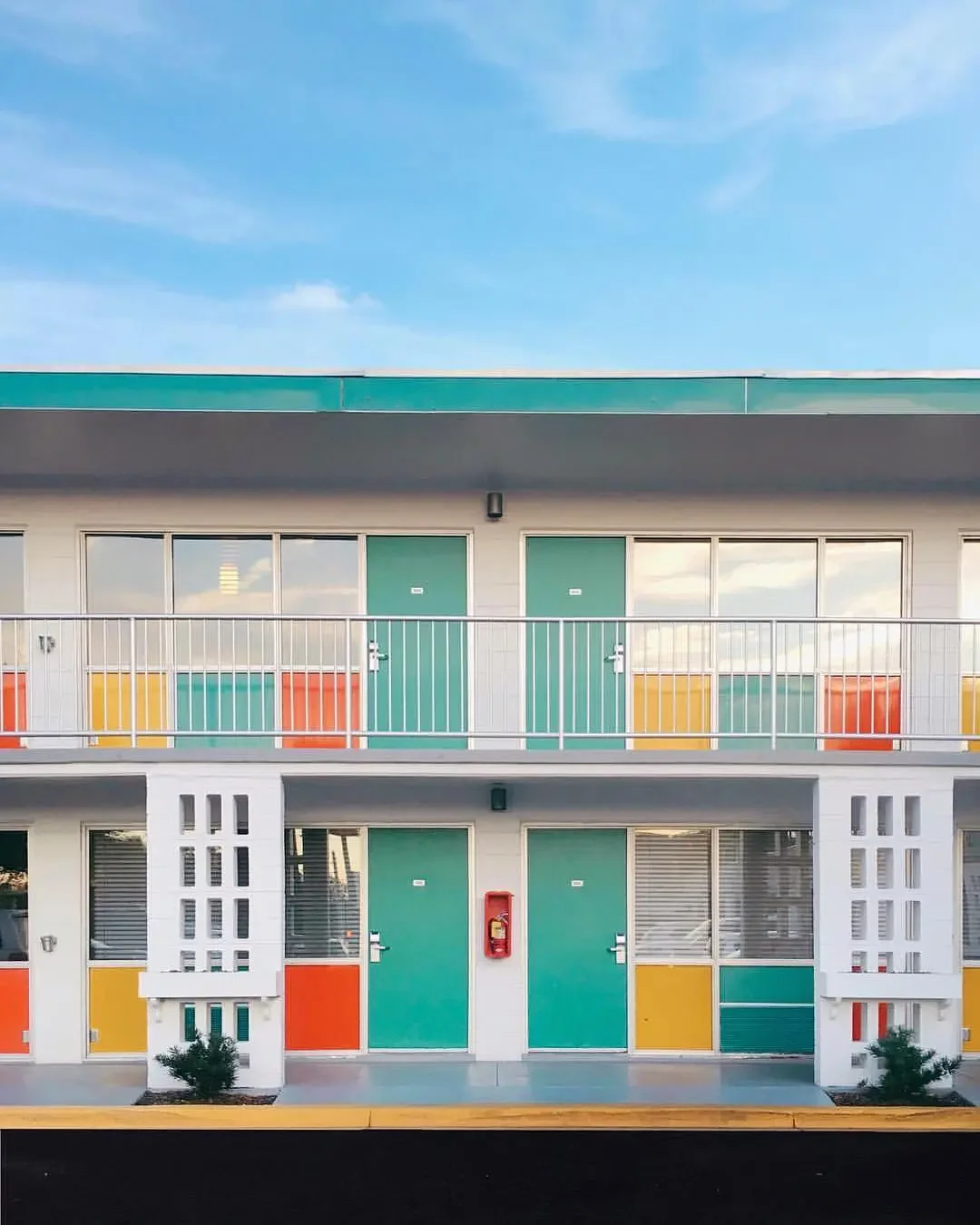 Two-story motel with pastel-colored doors and panels, white exterior walls, and a balcony with white railing, under a blue sky.