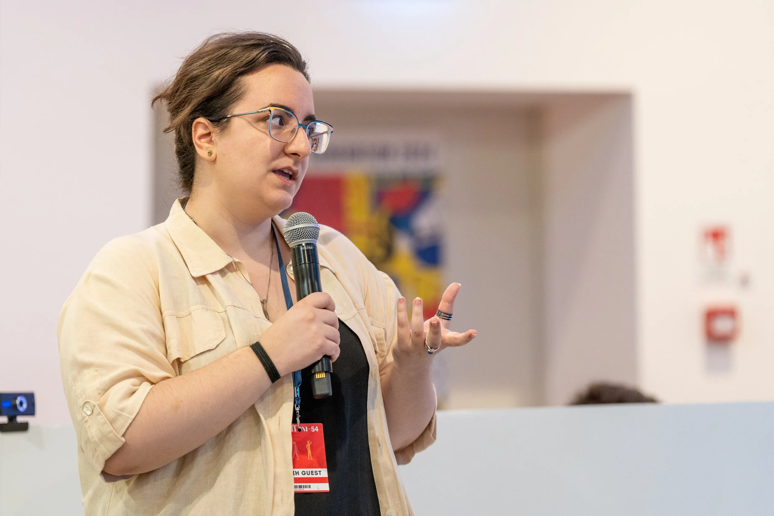 Donna parla con un microfono durante un evento, indossa occhiali e ha un badge con scritto 'Guest'.