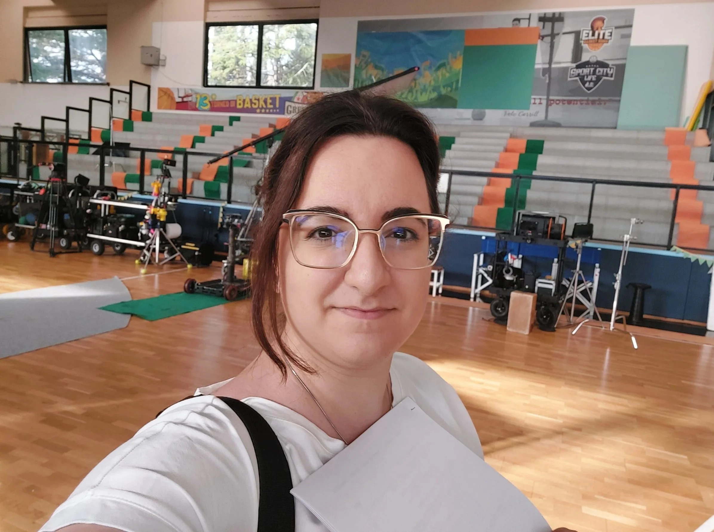 Ragazza che si prende un selfie in un centro sportivo o palestra, con gradinate colorate, attrezzature sportive e poster sul muro.