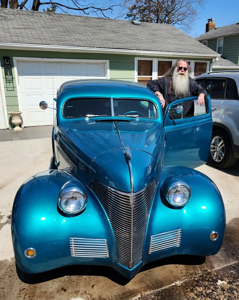 A vintage blue car parked in a driveway with a man with long hair and a beard leaning on the open driver's side door.