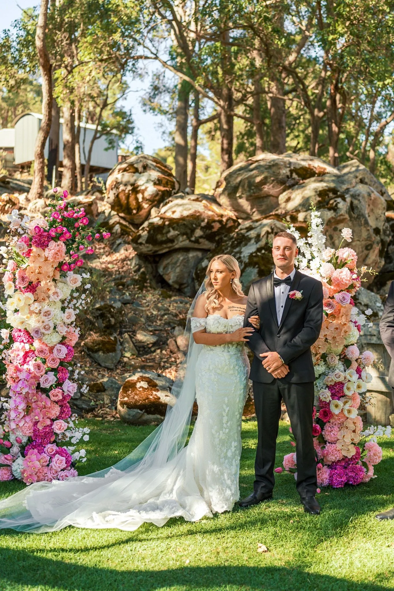 A bride and groom standing outdoors during a wedding ceremony, surrounded by pink and white floral arrangements, with trees and rocks in the background.