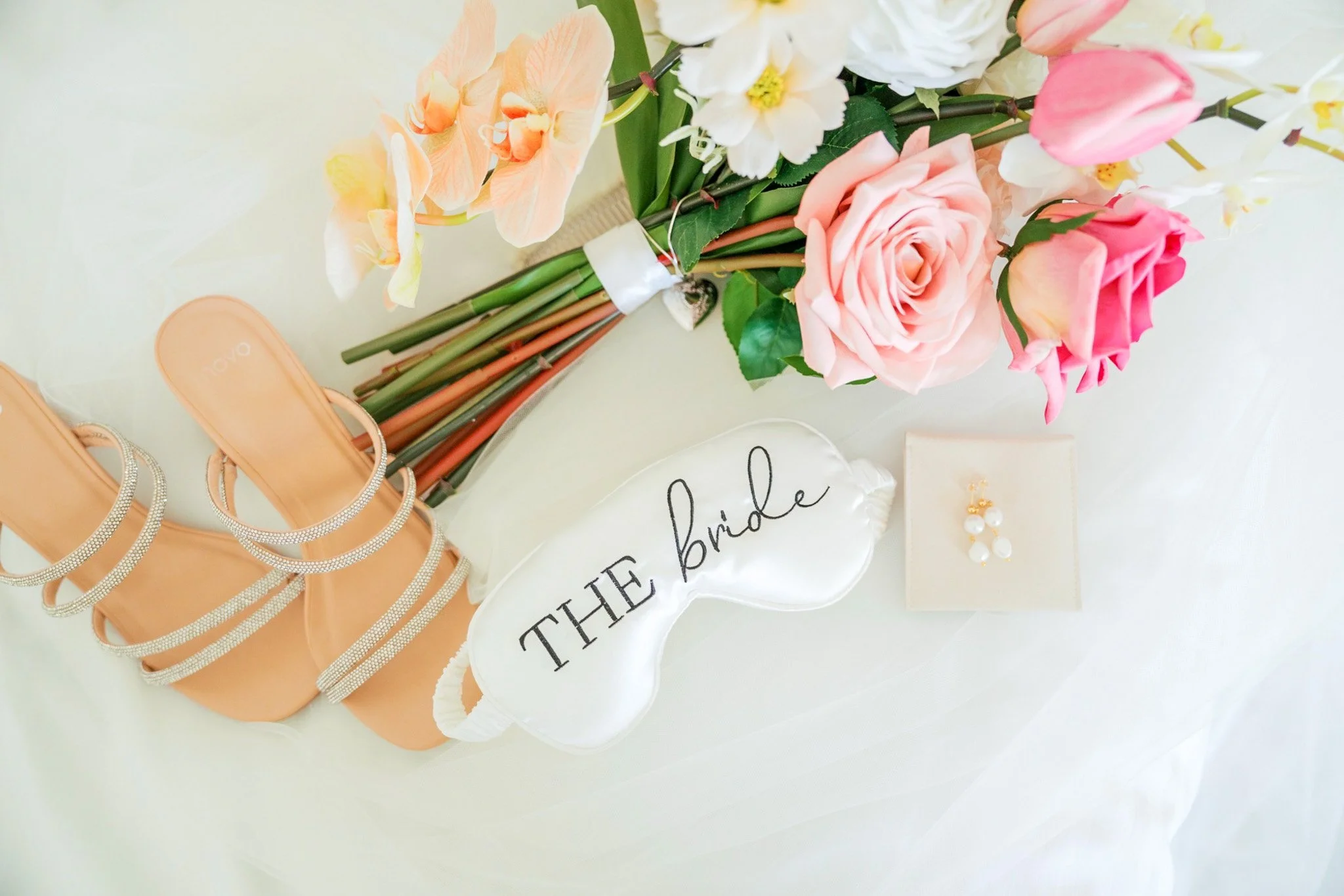A bouquet of pink, white, and peach flowers on a table next to blush-colored high-heeled shoes with rhinestone straps, a pair of pearl earrings, and a white sign with black script that reads "The bride".