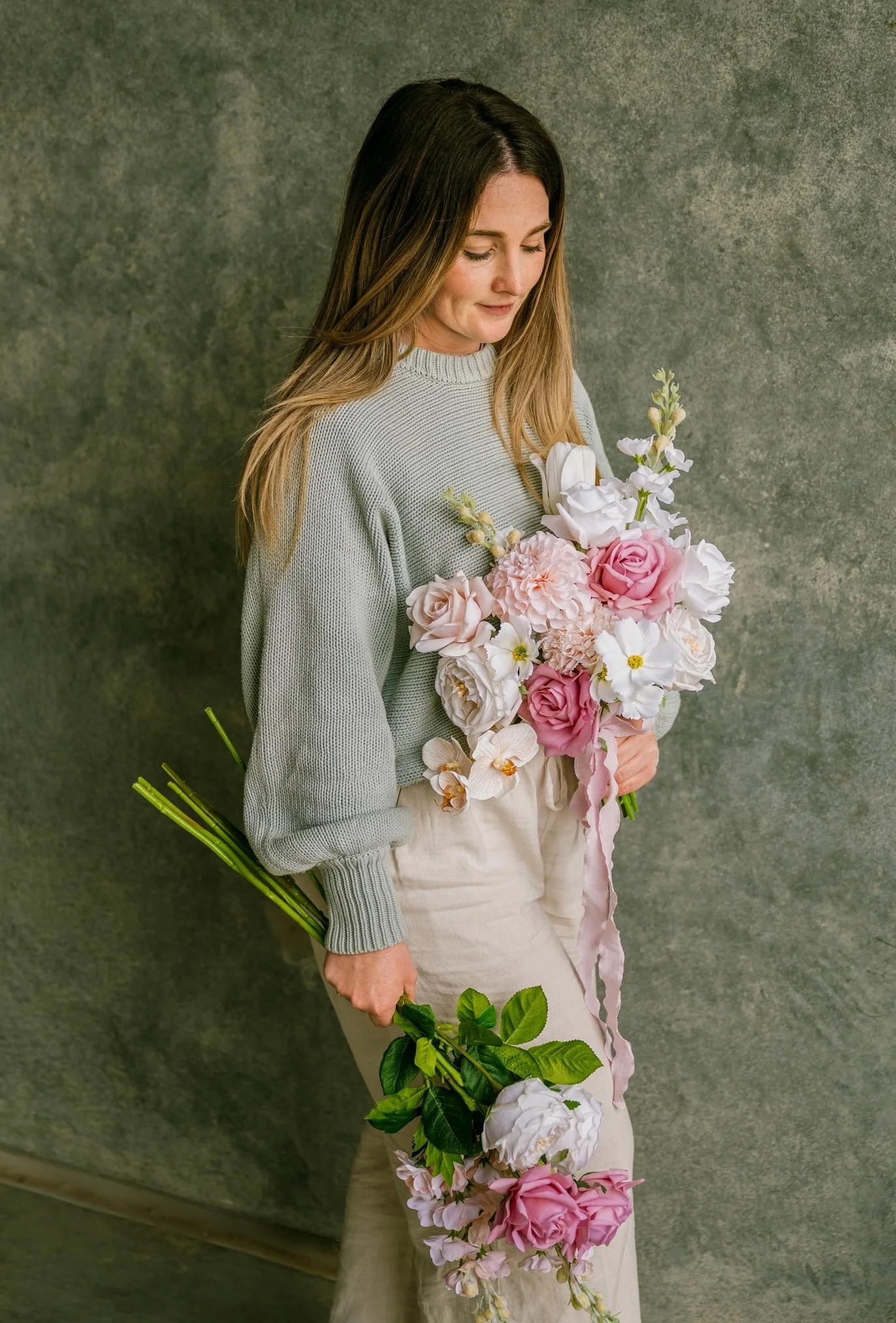 A woman with long brown hair wearing a light grey sweater, holding a large bouquet of pink and white flowers, standing against a green textured background.