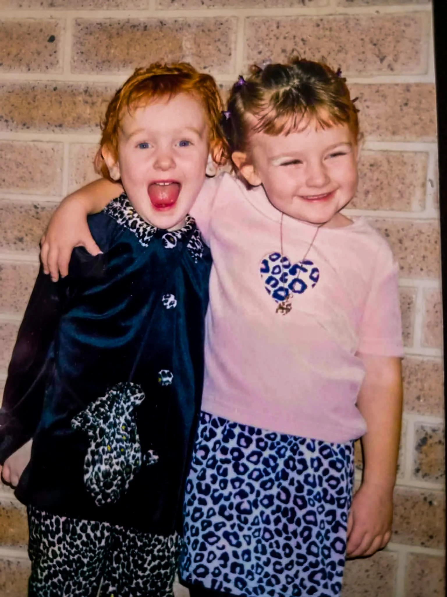 Two young girls with red hair, one with curly hair and another with straight hair, hugging each other and smiling. They are standing in front of a brick wall. The girl on the left is wearing a velvet dress with leopard print accents, and the girl on the right is wearing a pink shirt with a blue leopard print skirt and a heart-shaped necklace.