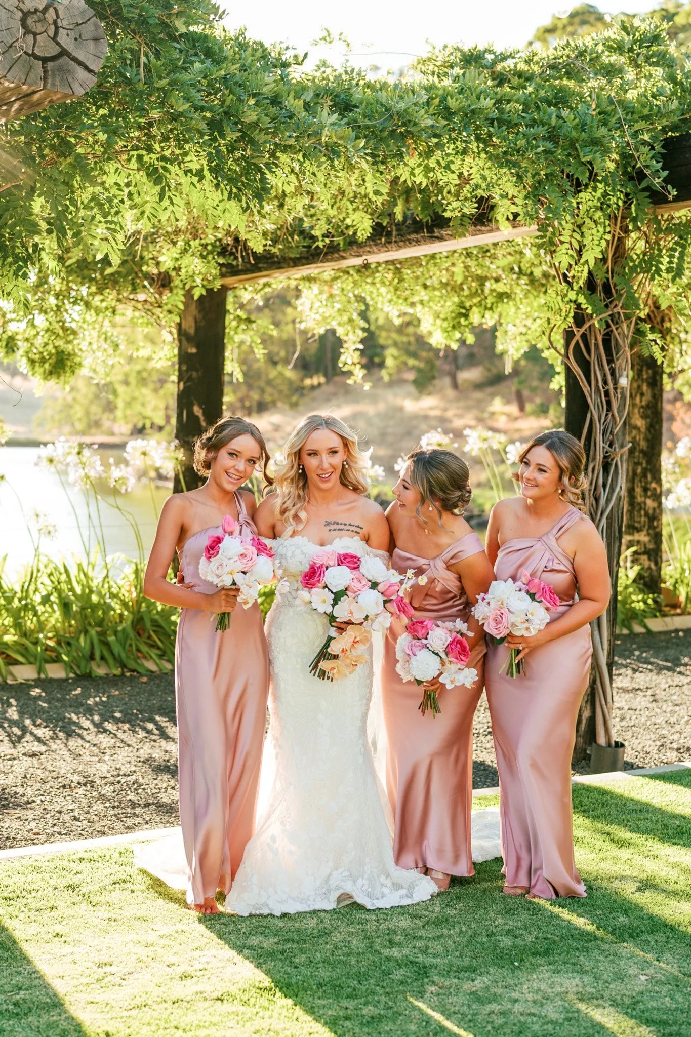 A bride in a white wedding dress holding a bouquet of pink and white roses, standing with four bridesmaids in blush pink dresses, outdoors under a wooden arch with green foliage, during a sunset.