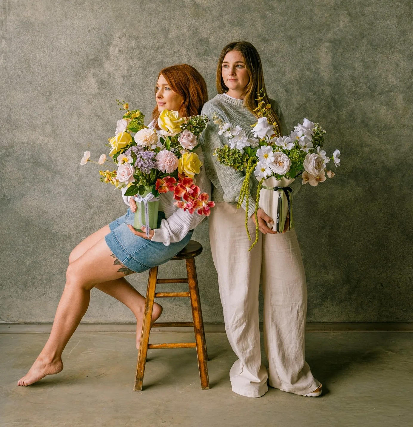 Two women holding large bouquets of flowers, one sitting on a wooden stool and one standing, against a plain grey background.