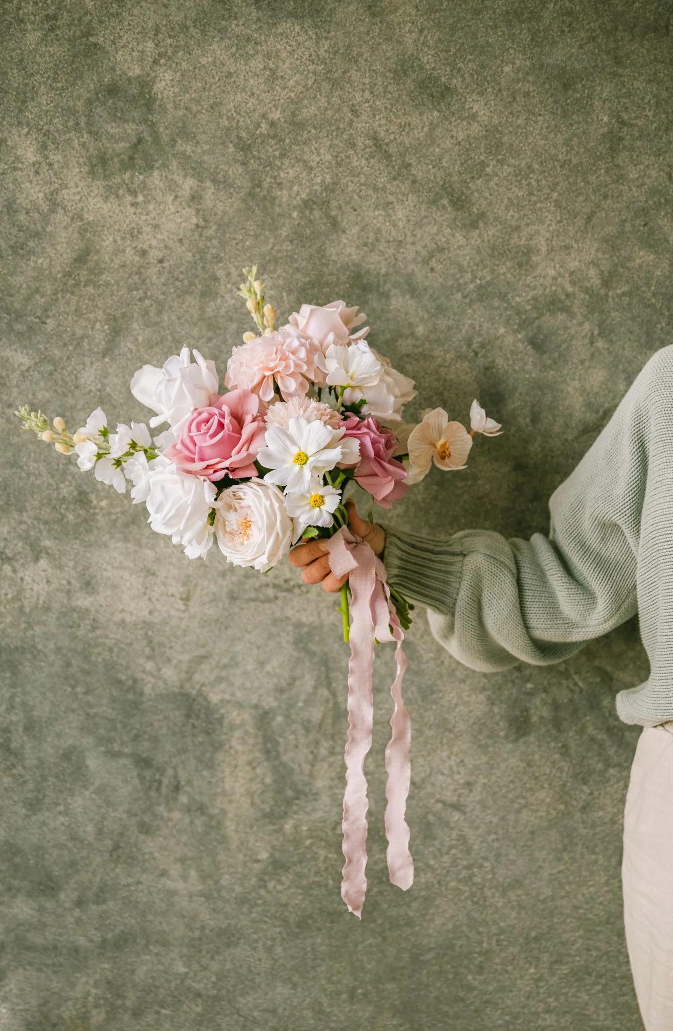 Person holding a bouquet of pink and white flowers with a pastel pink ribbon, wearing a gray sweater, against a textured gray background.