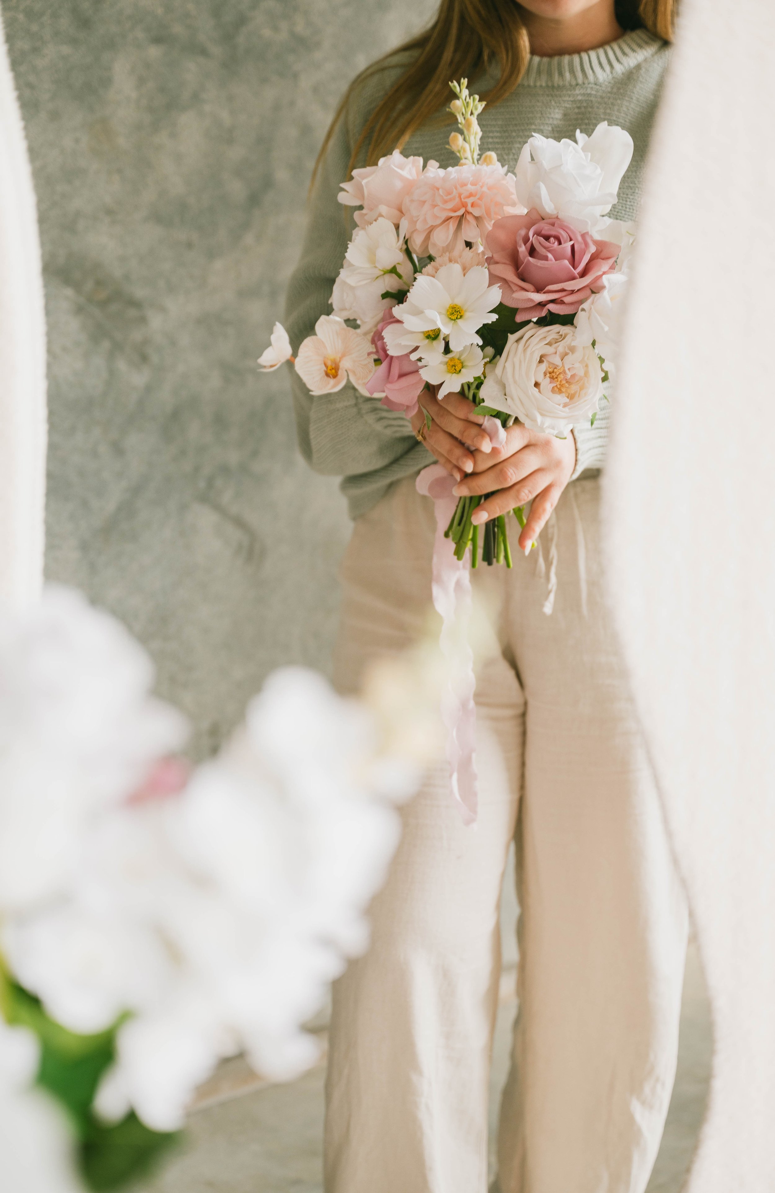 A woman holding a bouquet of various pink, white, and cream flowers, including roses, peonies, and daisies, in front of her chest, dressed in a light sweater and cream-colored pants.