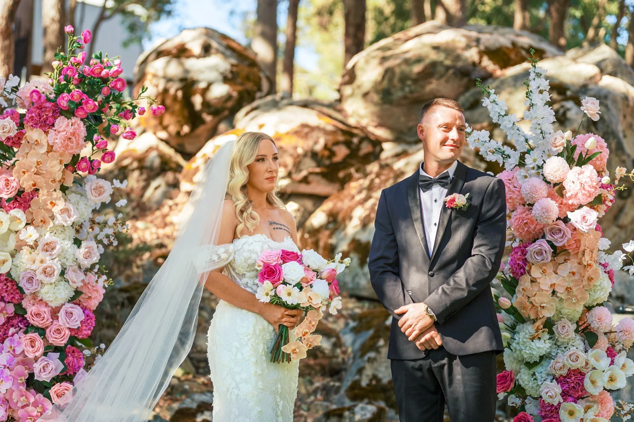 A bride and groom stand outdoors during their wedding ceremony, surrounded by colorful pink and white flowers in an outdoor setting with trees.