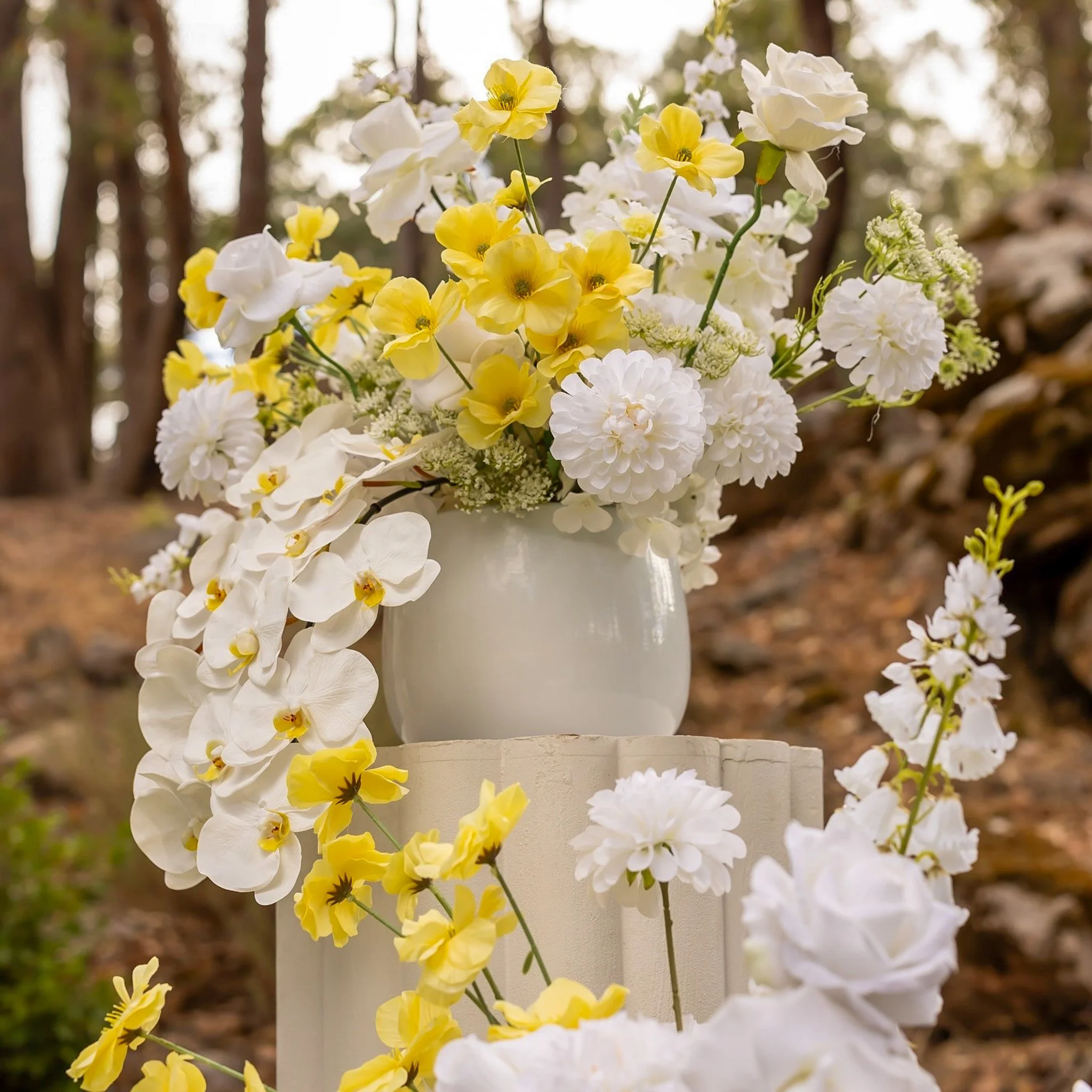 A bouquet of white and yellow flowers arranged in a white vase outdoors with a blurred background of trees.