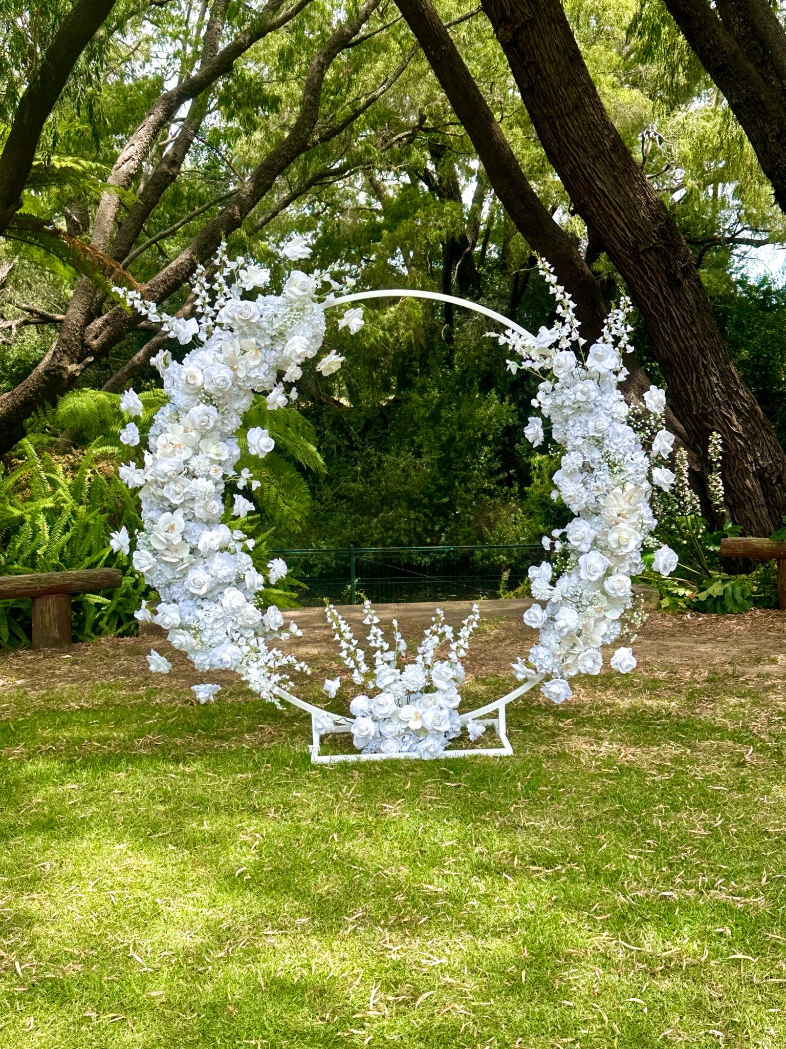 A circular wedding arch decorated with white flowers, set in a lush outdoor garden with green trees and grass.