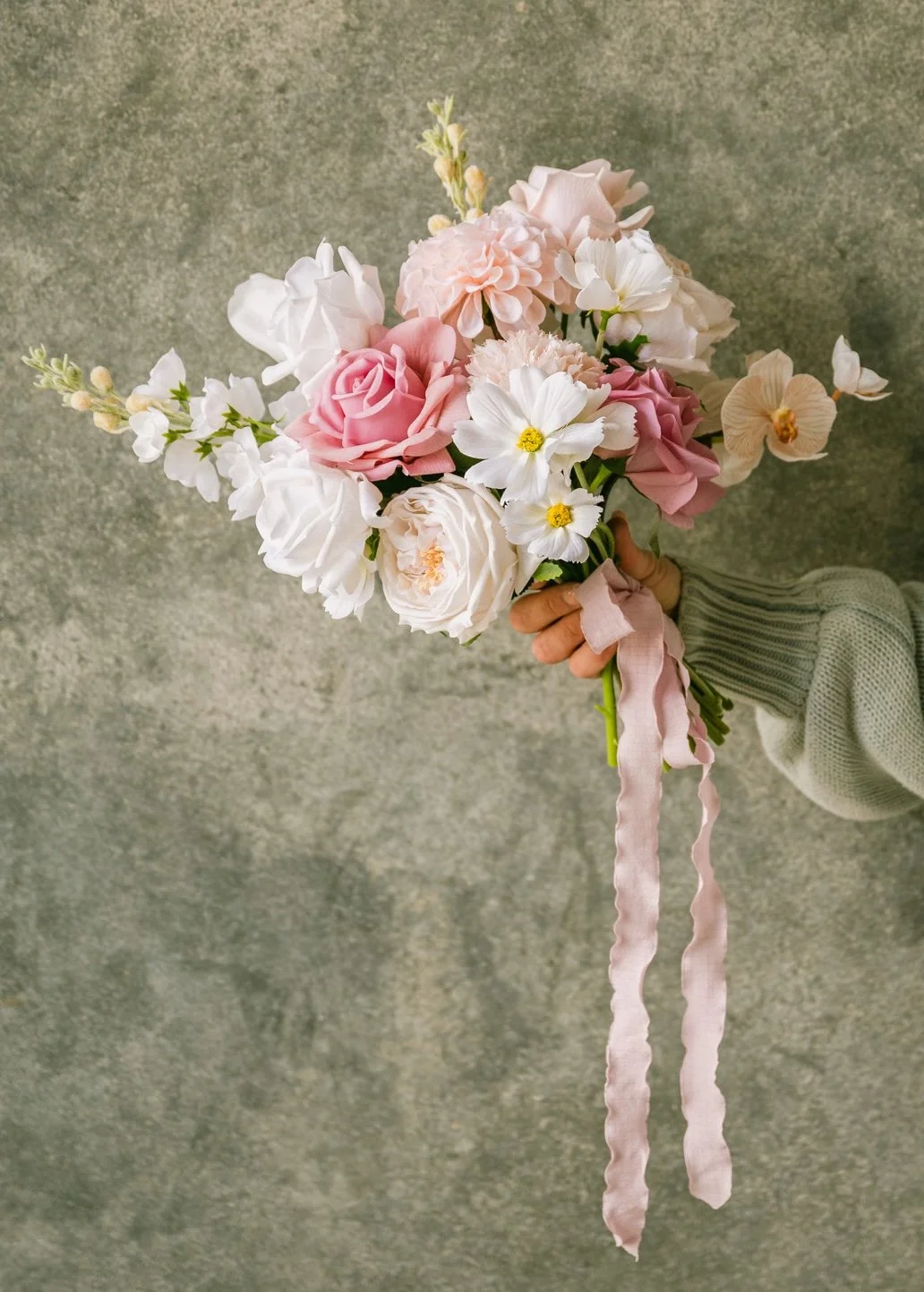A hand holding a bouquet of pink, white, and cream flowers with a pink ribbon against a gray textured background.