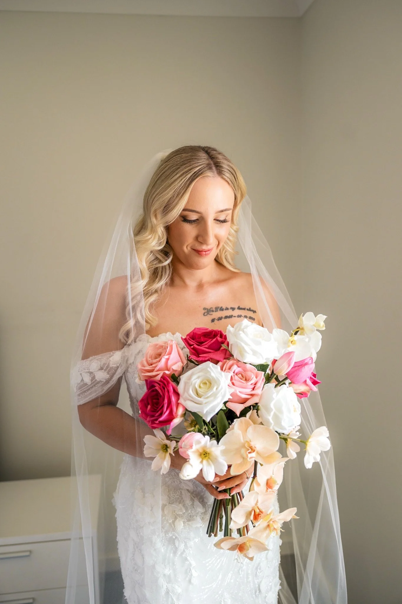 A bride with blonde hair in loose curls, wearing a white lace wedding dress and veil, holding a bouquet of pink, white, and cream roses and orchids, looking down with a gentle smile.
