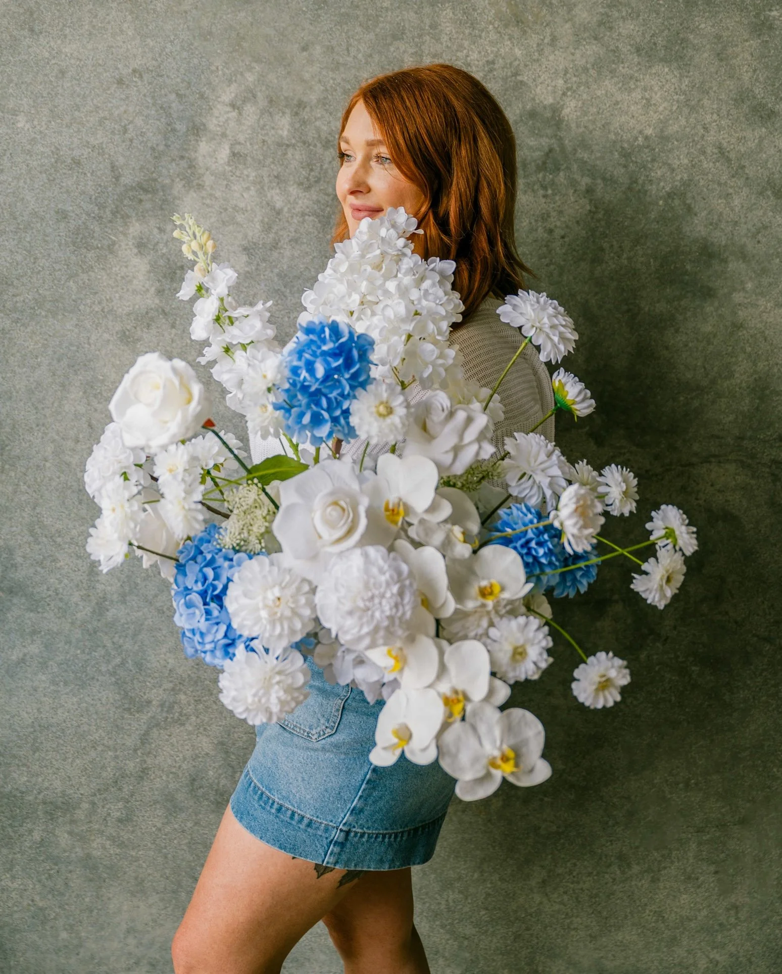 A woman with red hair wearing a gray shirt and denim shorts, holding a large bouquet of white and blue flowers, standing against a gray textured wall.