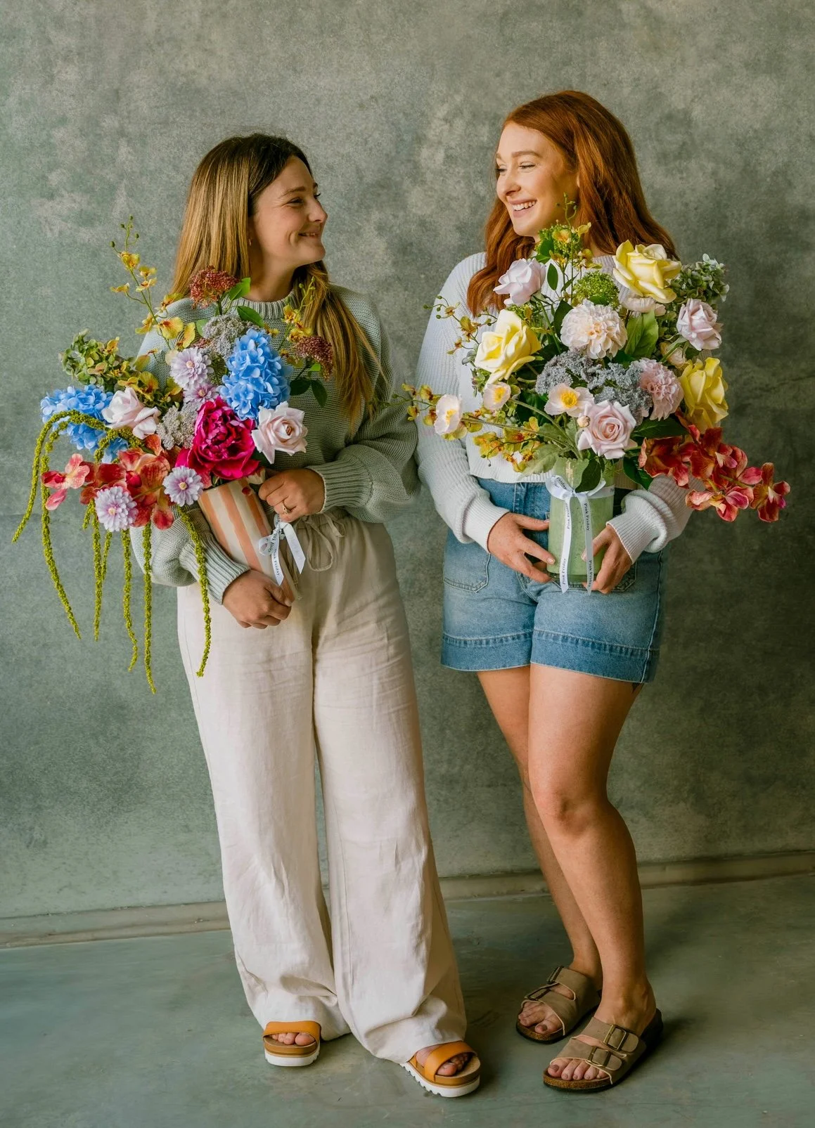 Two women standing side by side, each holding a large bouquet of colorful flowers. They are smiling and looking at each other, with a plain gray background behind them.