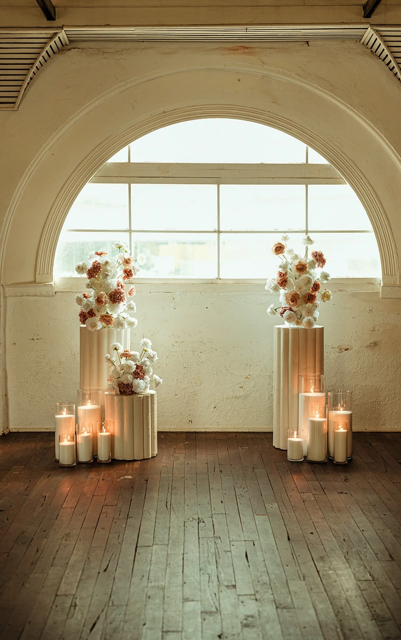 Decorative floral arrangements with candles on wooden pedestals set against a large window with arched molding and a rustic wooden floor.