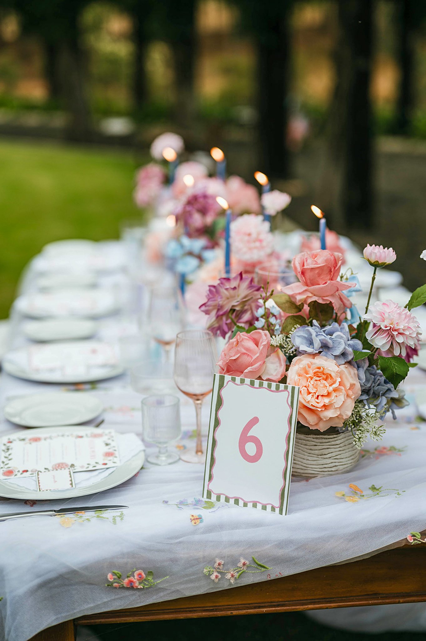A decorated outdoor table with a floral centerpiece, candles, table setting, and a table number card with the number 6. The table is set for a celebration or special event.
