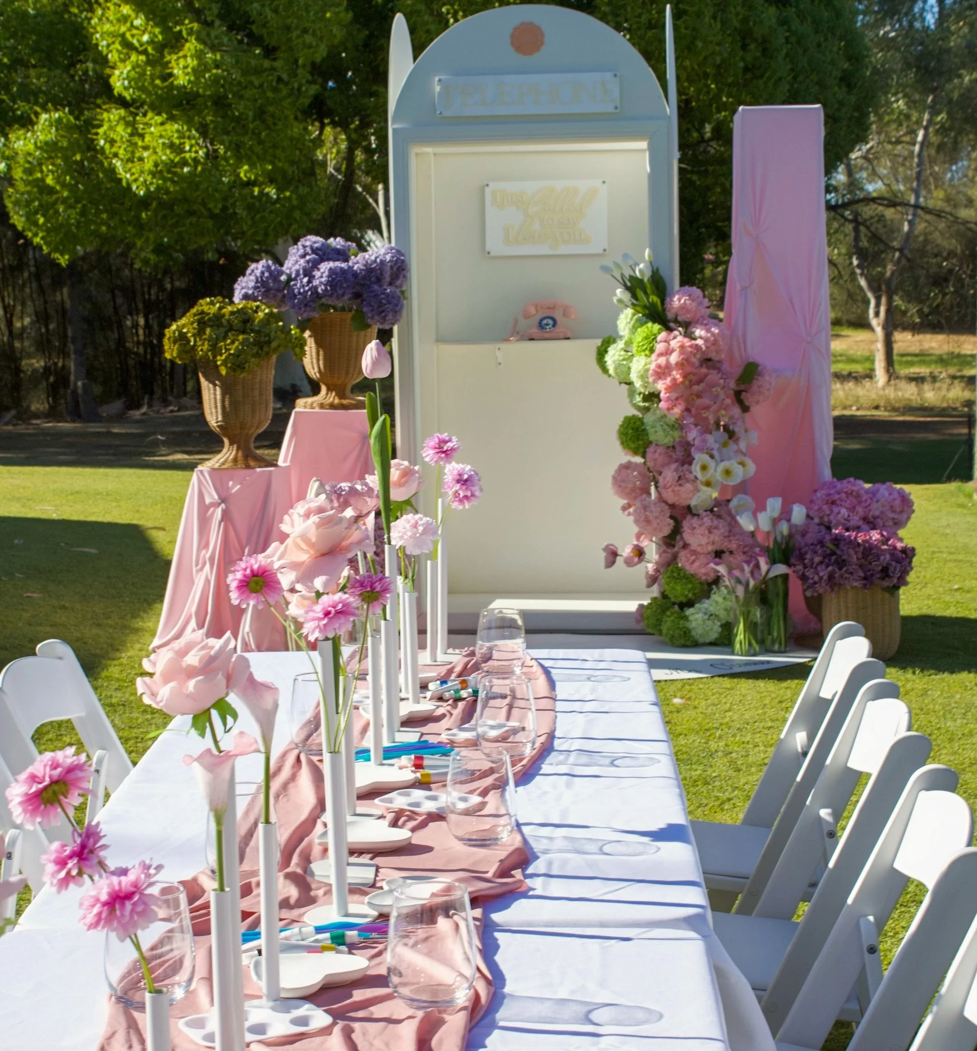 Decorated outdoor table for a celebration or party with pink and purple flowers, pink and white tablecloth, chairs, and a floral backdrop, set in a green garden or park.