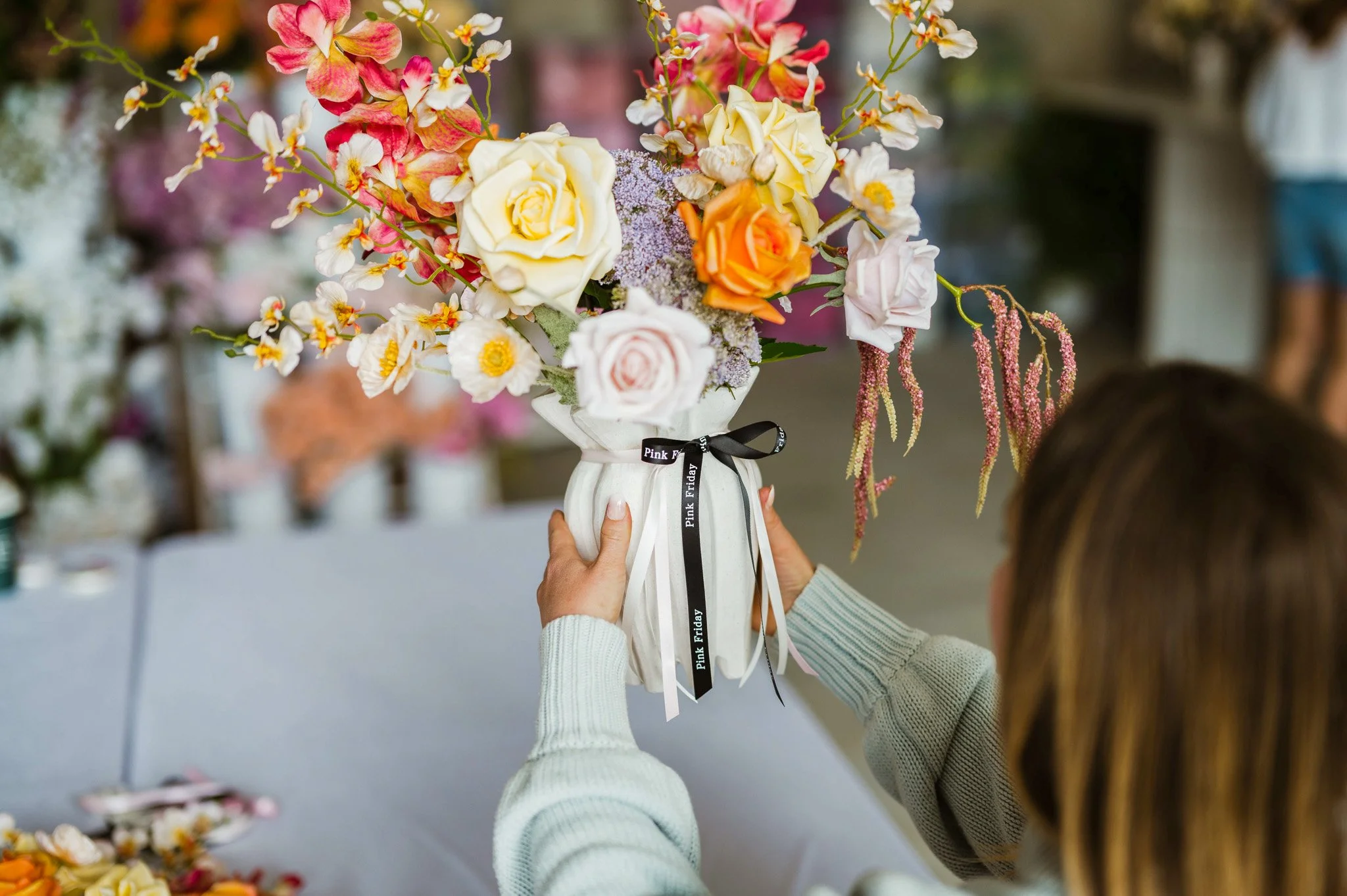 Person holding a white vase filled with various colorful flowers, including roses, orchids, and other blossoms, with a blurred background of more flowers.