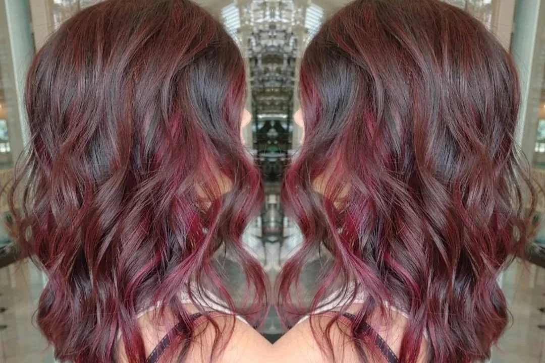 Mirror image of a woman with long, wavy, dark burgundy hair in a salon setting.