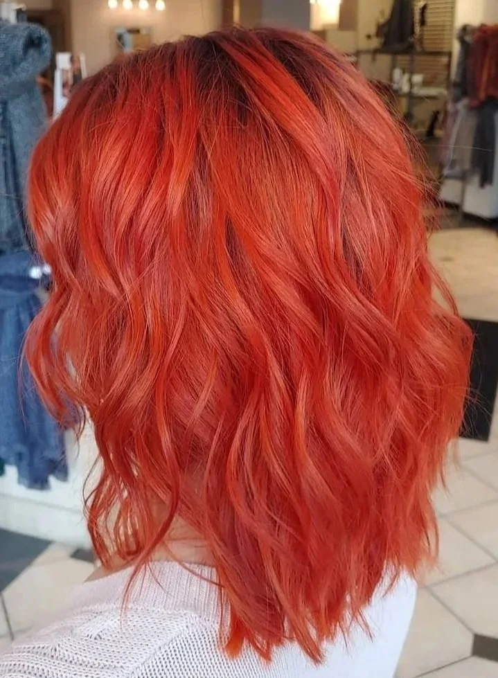 Woman with shoulder-length, wavy, bright orange-red hair in a salon or store setting.