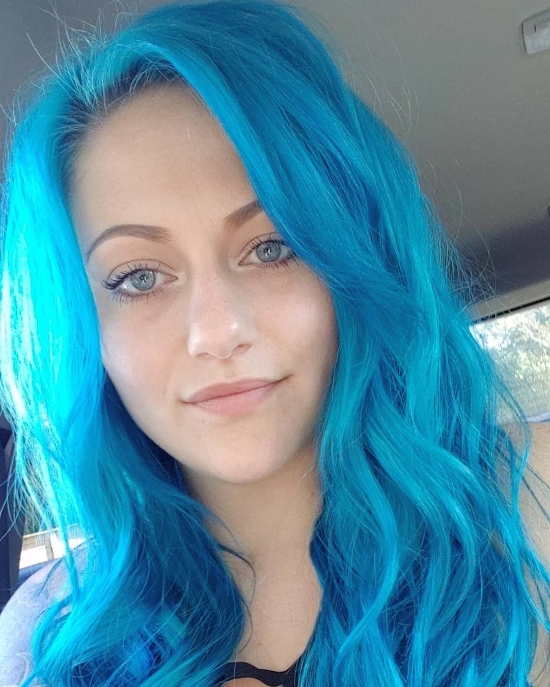 Close-up of a young woman with bright blue wavy hair and light blue eyes, smiling softly at the camera inside a vehicle.