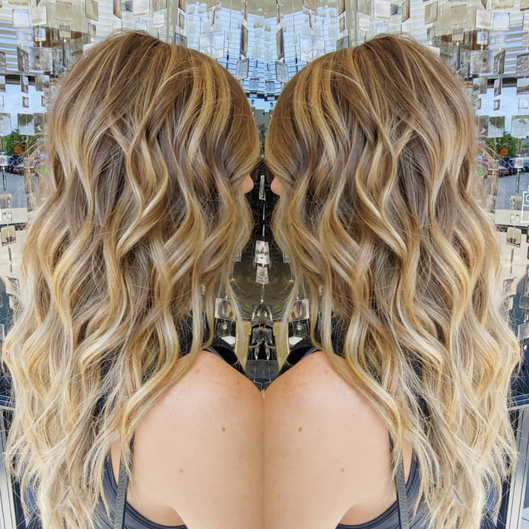 A mirror behind a woman with long, wavy blonde hair, showing her reflection from the back in a store or salon setting.