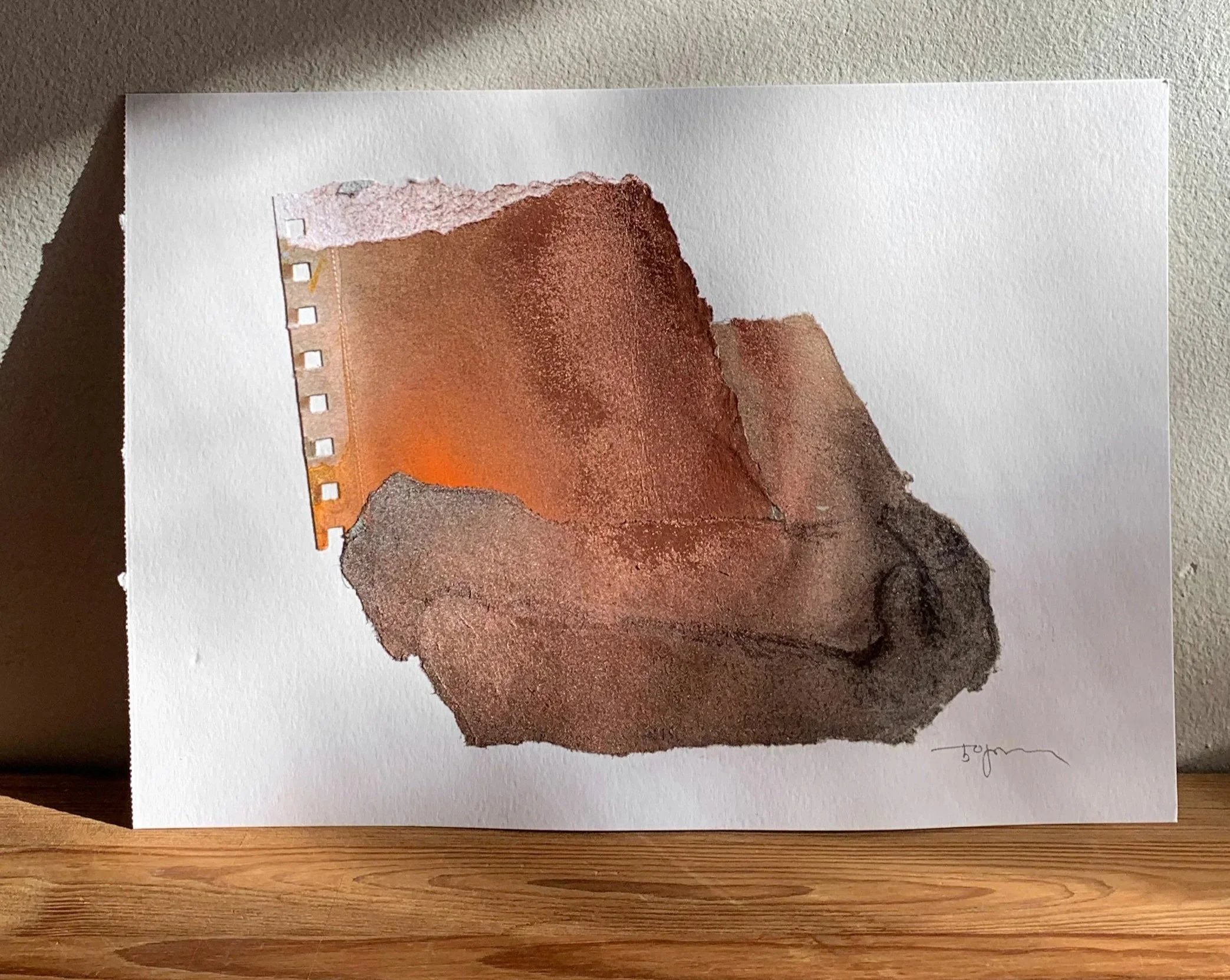 Abstract watercolor painting on white paper depicting a combination of textured, earthy-toned shapes with a small section of orange near the top left. The artist's signature is visible in the bottom right corner.