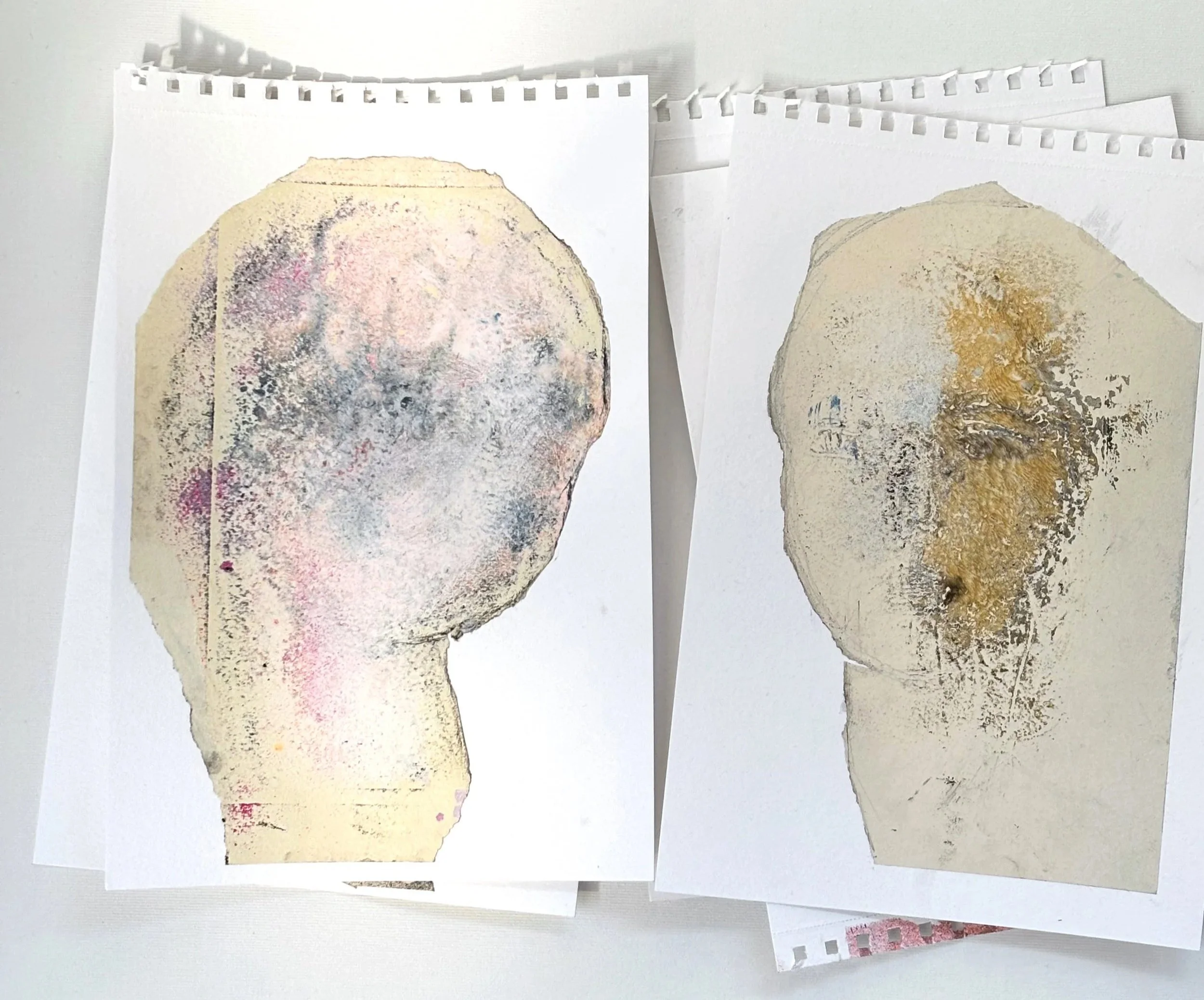 Several pieces of paper with abstract, textured prints of human head silhouettes, one with pink and gray colors and the other with brown and gray tones.
