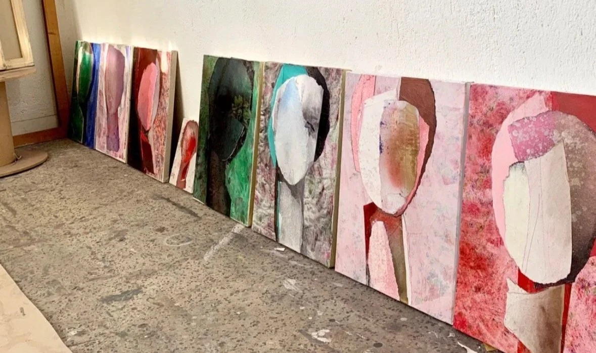 A series of colorful abstract paintings leaning against a white wall in an art studio or gallery space.