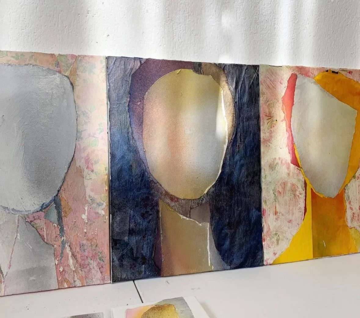 Abstract watercolor paintings on paper, displayed against a white wall, featuring organic shapes and muted colors.