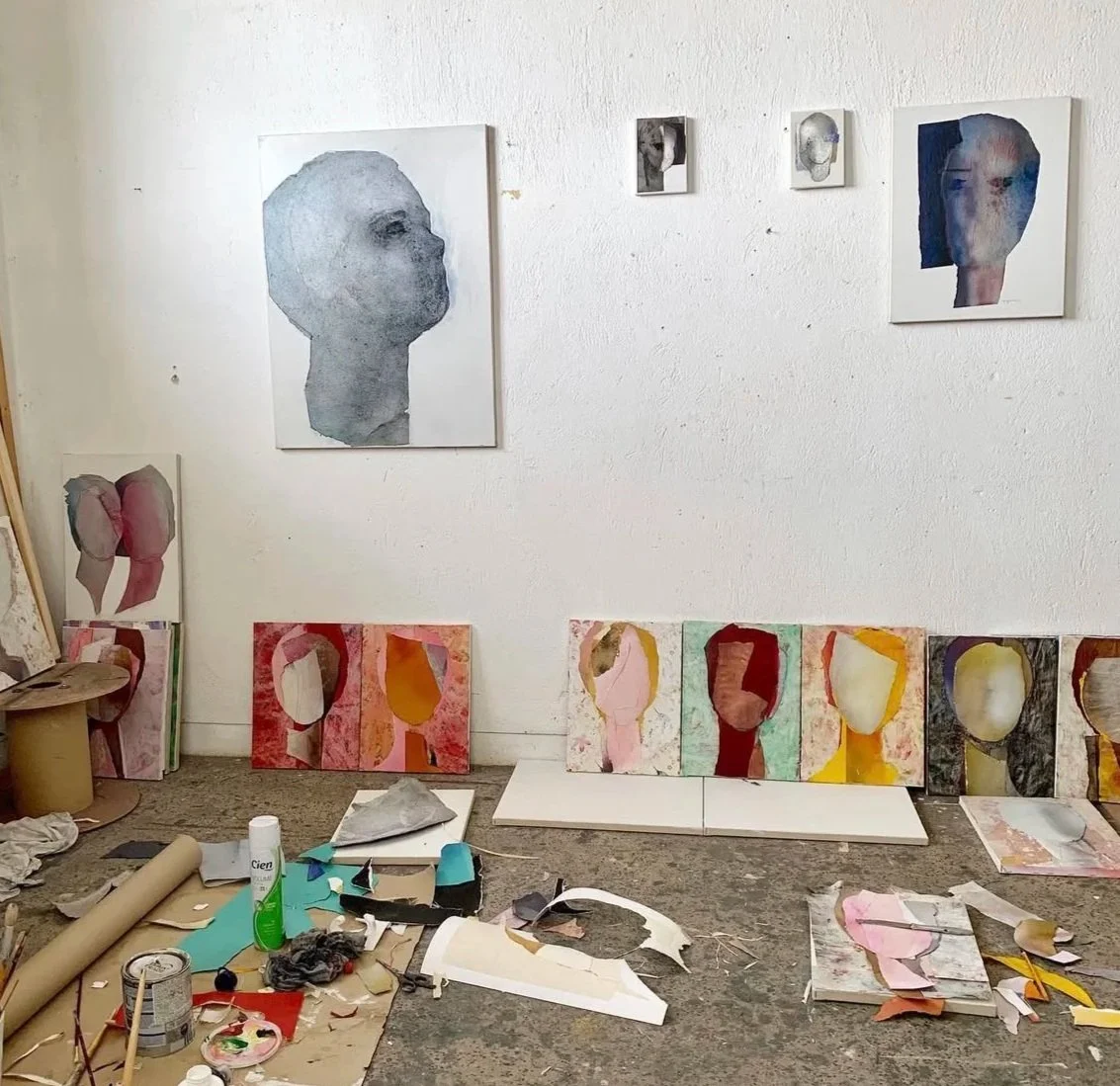 Art studio walls with paintings of abstract human faces, some finished and some in progress, surrounded by art supplies, paints, brushes, and canvases on the table and floor.