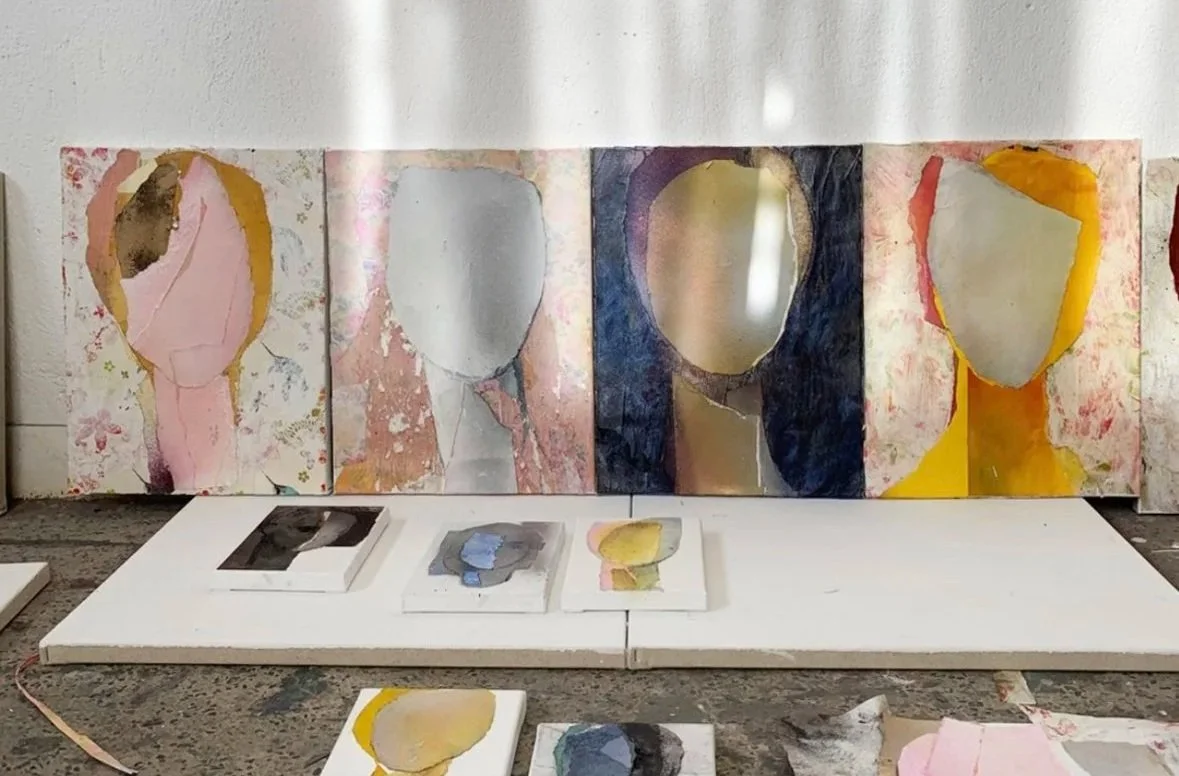 Art display with abstract portrait paintings featuring oval shapes for faces, in pink, gray, blue, and yellow colors, arranged on a white shelf and table.