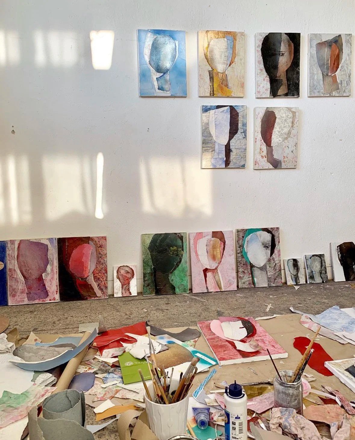 A wall with multiple abstract portrait paintings, some are finished and some are in progress, with art supplies scattered on the floor below.