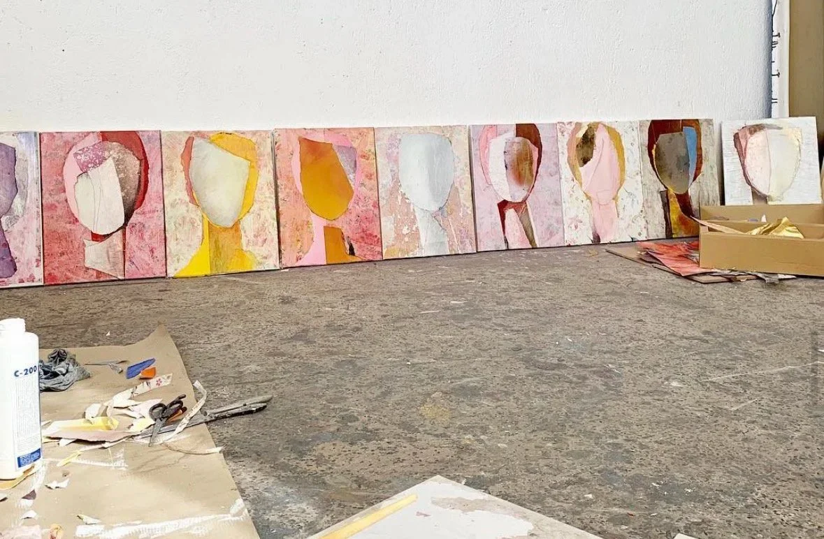 A row of abstract portrait paintings leaning against a white wall, with art supplies like paper, scissors, glue, and paint on the floor in the foreground.