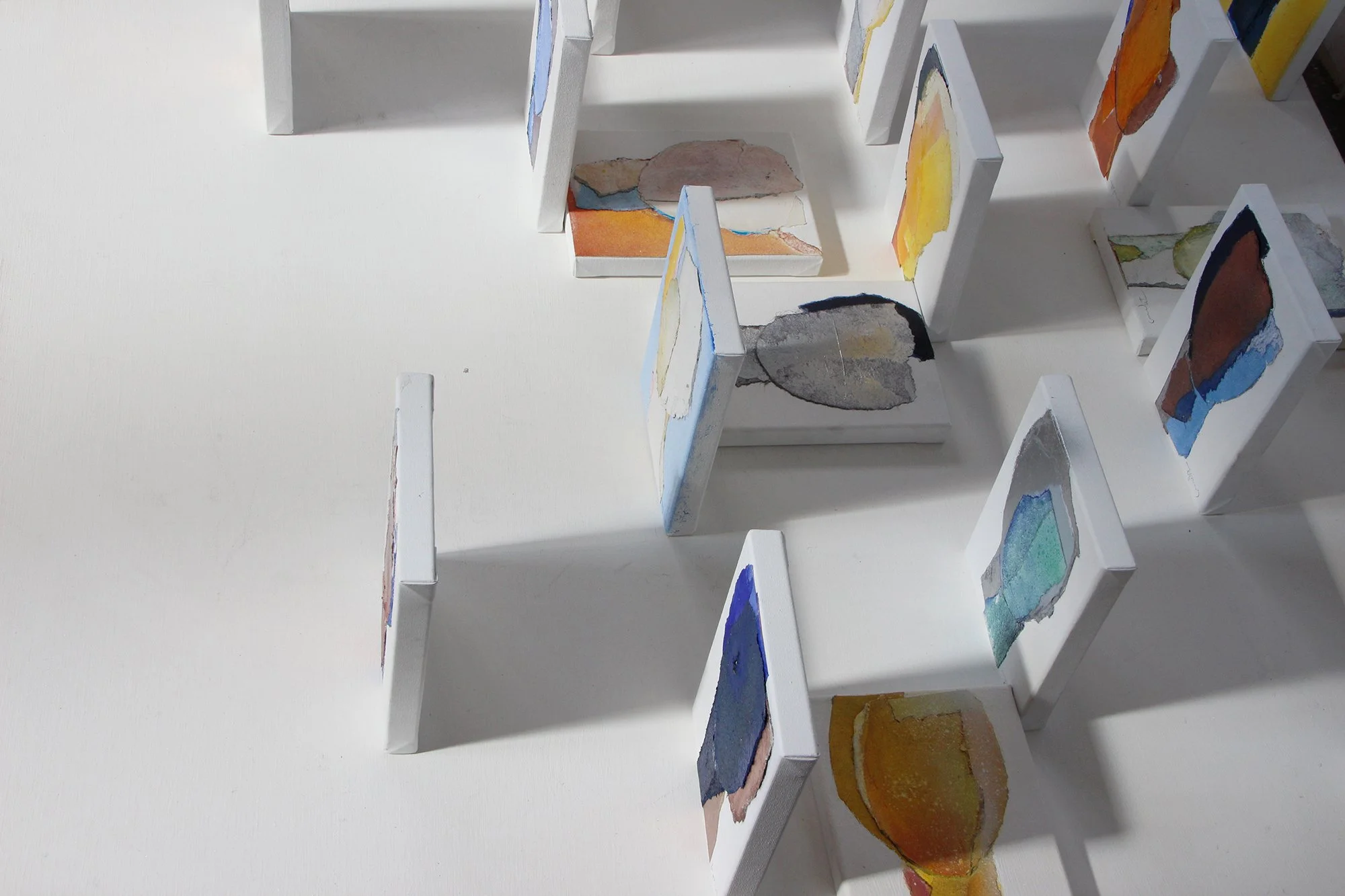 Miniature art display with watercolor paintings on small vertical and horizontal surfaces, arranged in a grid pattern on a white background.