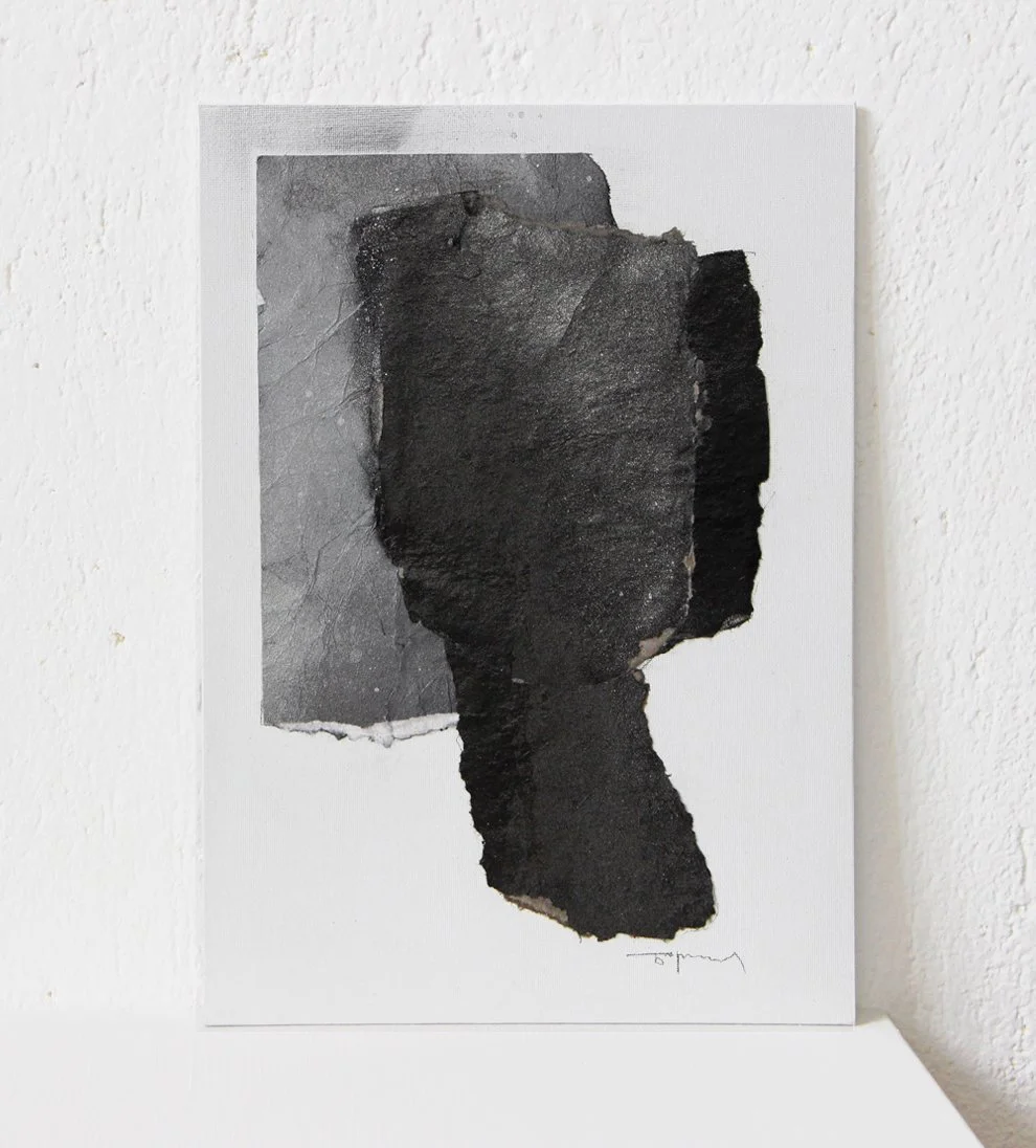 Abstract black and gray textured collage on a white canvas leaning against a white wall.