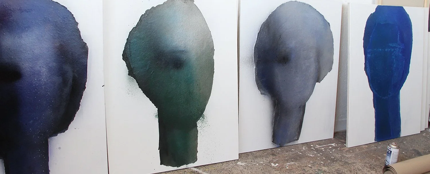 Four abstract watercolor paintings on large canvases, each depicting a stylized face in dark colors, leaning against a wall in an art studio.