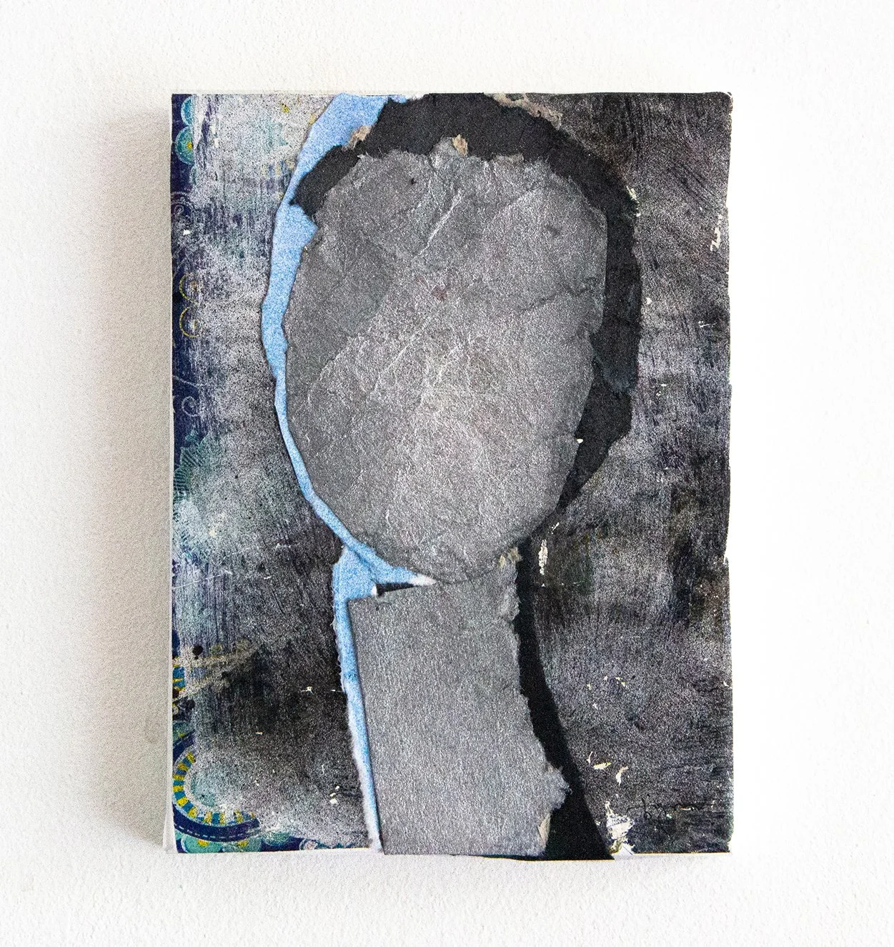 A piece of artwork featuring multiple layers of torn paper with textured gray, black, and blue colors, arranged in an abstract composition.