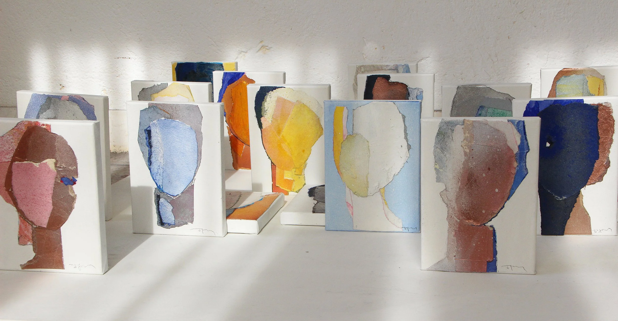 Artwork featuring abstract, minimalist portraits of faces, painted in watercolor on white canvases arranged in groups on a white surface against a white wall.