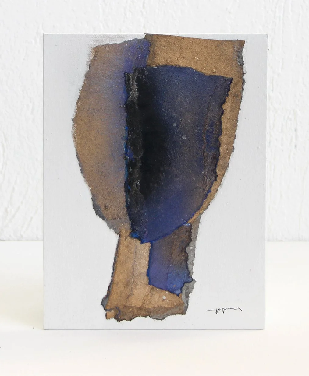 An abstract mixed media artwork on paper, featuring a shape resembling a shield or a face, with blue, black, and brown colors.