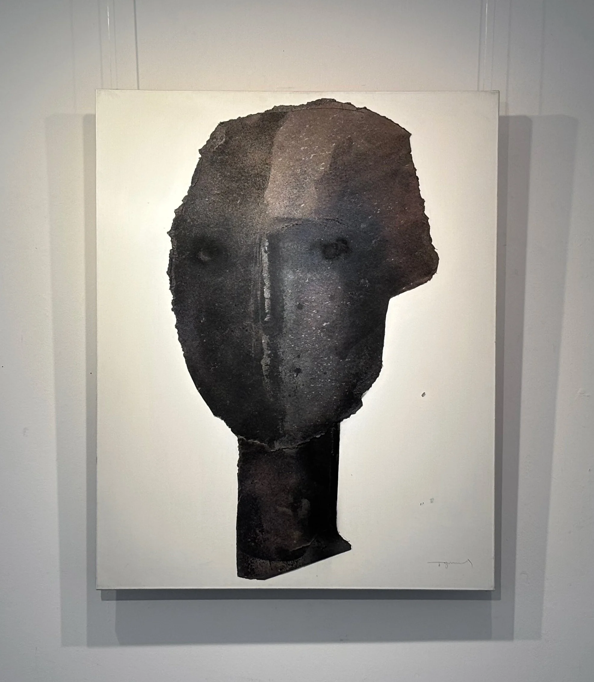 A painting of a humanoid face with a textured, dark surface and pale eyes, mounted on a white wall.