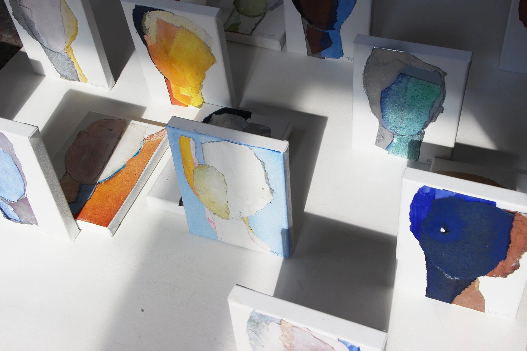 Multiple small abstract watercolor paintings with torn paper edges, featuring various shapes and colors, displayed on a white surface with shadows.