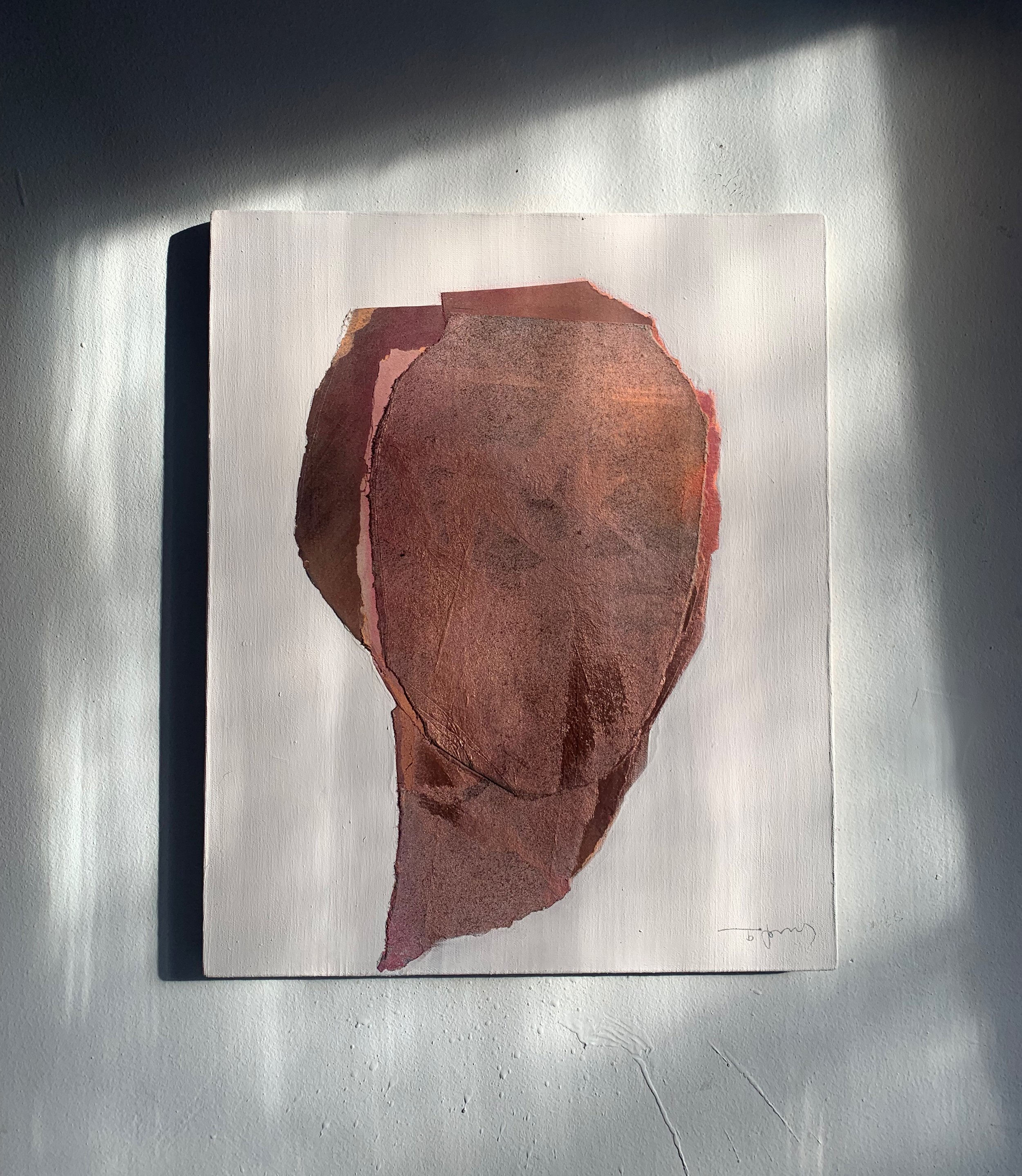 Abstract artwork of a face in earthy tones, mounted on a white wall with sunlight casting shadows.